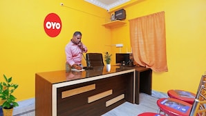 Uma mesa de recepção com um homem atrás dela, uma planta sobre a mesa e uma parede amarela com o logo 'OYO'.