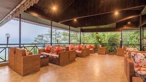 A covered outdoor seating area with wicker furniture and orange cushions, overlooking a lush green landscape. - Udaan Hotels Alpine Resort, Gangtok (Gangtok)