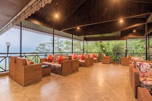 A covered outdoor seating area with wicker furniture and orange cushions, overlooking a lush green landscape.