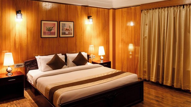 A hotel room with a wooden paneled wall, a bed with white bedding and brown pillows, two bedside lamps, and a window with curtains. - Udaan Hotels Alpine Resort, Gangtok (Gangtok)