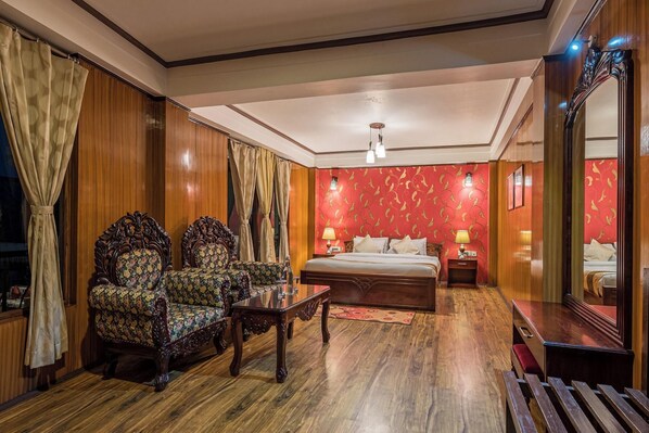Ein Hotelzimmer mit einem großen Bett, zwei verzierten Sesseln, einem Holztischchen, einem Nachttisch mit Lampen und einem Spiegel.