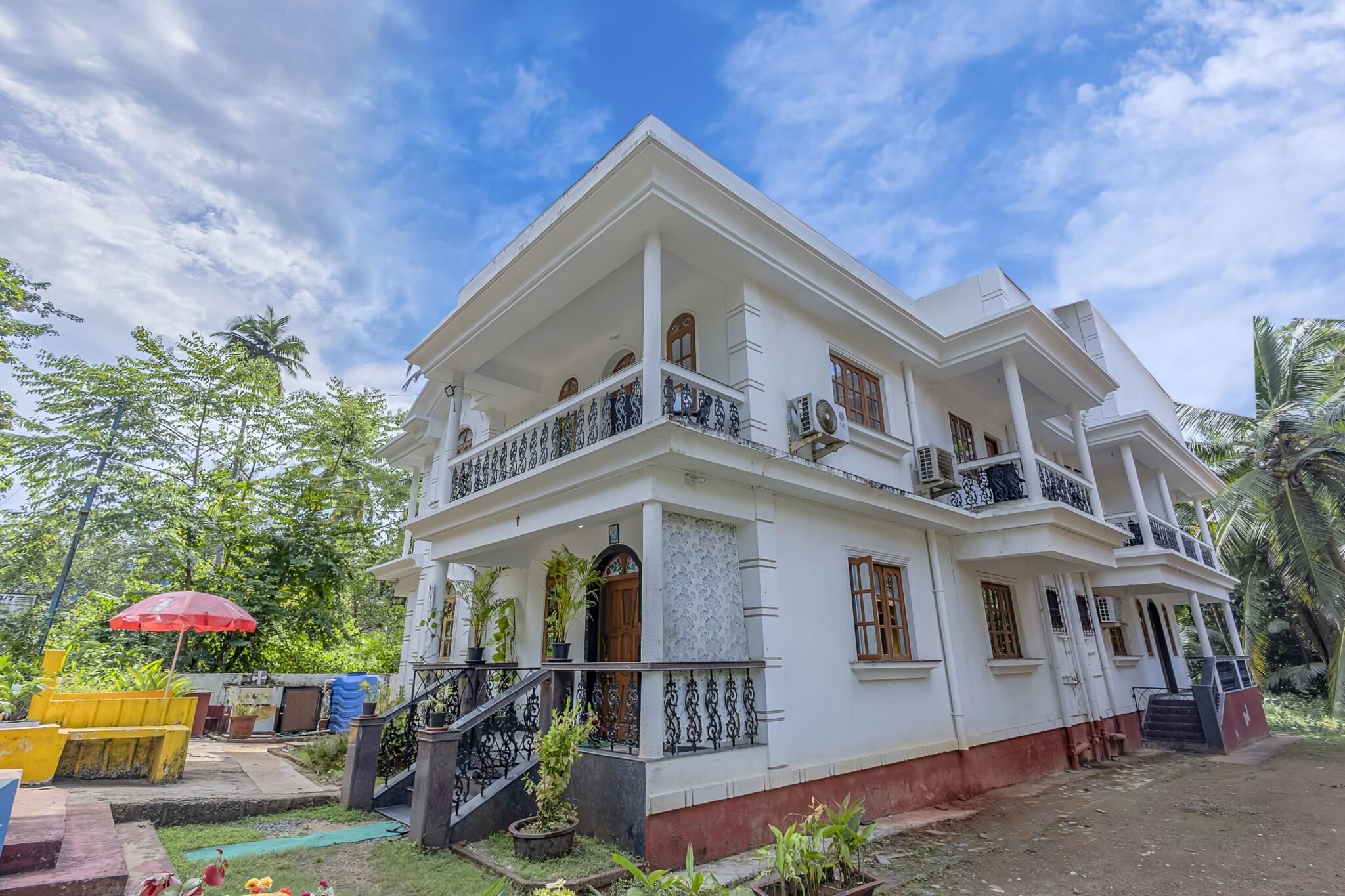 A two-story white building with balconies, a staircase, and a garden area with plants and a red umbrella.