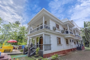A two-story white building with balconies, a staircase, and a garden area with plants and a red umbrella. - The Sequeira By Madhu And Sons (Majorda)