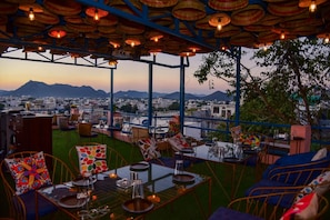 An outdoor seating area with tables and chairs, a view of a cityscape, and a mountain in the background. - Casa By The Lake (Udaipur)