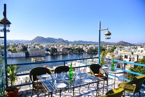 A terrace with a table set for dining, overlooking a river and cityscape with mountains in the background. - Casa By The Lake (Udaipur)