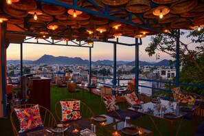 An outdoor seating area with tables and chairs, a view of a cityscape, and a mountain in the background. - Casa By The Lake (Udaipur)
