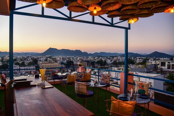 A rooftop terrace with tables and chairs, a view of a cityscape and mountains in the background. - Casa By The Lake (Udaipur)
