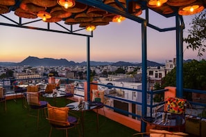 A terrace with tables and chairs, a view of a cityscape, and mountains in the background. - Casa By The Lake (Udaipur)