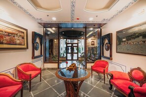 A room with a glass-top table, red upholstered chairs, and ornate wall decorations. - The Chronicles Hotel (Udaipur)