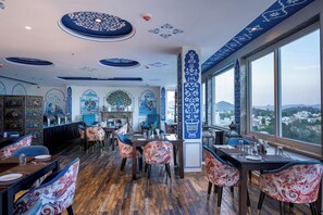 A restaurant with a high ceiling decorated with blue patterns, wooden flooring, and a view of the city through large windows. - The Chronicles Hotel (Udaipur)