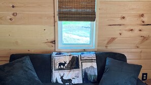A wooden cabin interior with a dark green sofa, a window with a view, and a patterned rug. - Cabin # 3. Sammy’s Deer Den  (Lima)