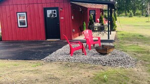 A red barn with a black door, a covered porch, and a gravel area with red chairs. - Cabin # 3. Sammy’s Deer Den  (Lima)