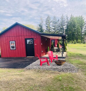 A red barn with a black door, a covered porch, and a gravel area with red chairs.
