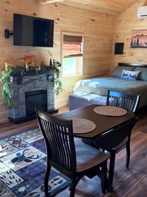 A cozy cabin interior with a fireplace, a television mounted on the wall, a dining table with chairs, and a sofa in the background.