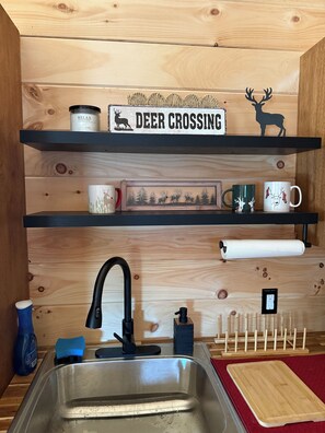 A kitchen with a sink, a coffee maker, a mug, a framed picture, and a shelf with a deer silhouette and a sign.