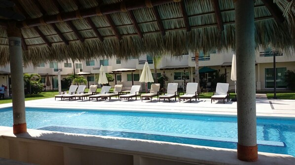 A pool area with lounge chairs and umbrellas, a thatched roof, and a building with multiple windows in the background.
