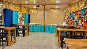 Ein Restaurant mit Holztischen und -stühlen, einem blauen Vorhang im Hintergrund und einer Wand mit Regalen voller Gegenstände.