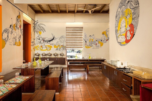A restaurant with wooden benches, a long dining table, and murals on the walls. - Colony Artotel (Kochi)