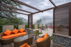 An outdoor patio with wicker furniture, a glass-top table, and a view of a mountainous landscape. - Hotel La Eden (Dehradun)