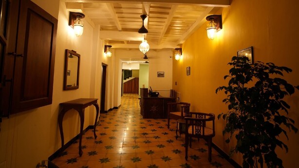 A hallway with wooden flooring, a wooden door, a mirror, a table, chairs, a plant, and wall-mounted lights.