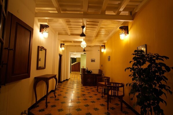 A hallway with wooden flooring, a wooden door, a mirror, a table, chairs, a plant, and wall-mounted lights.