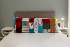 Een bed met een wit laken, een grijze hoofdbord en twee sierkussens met lappendecor.