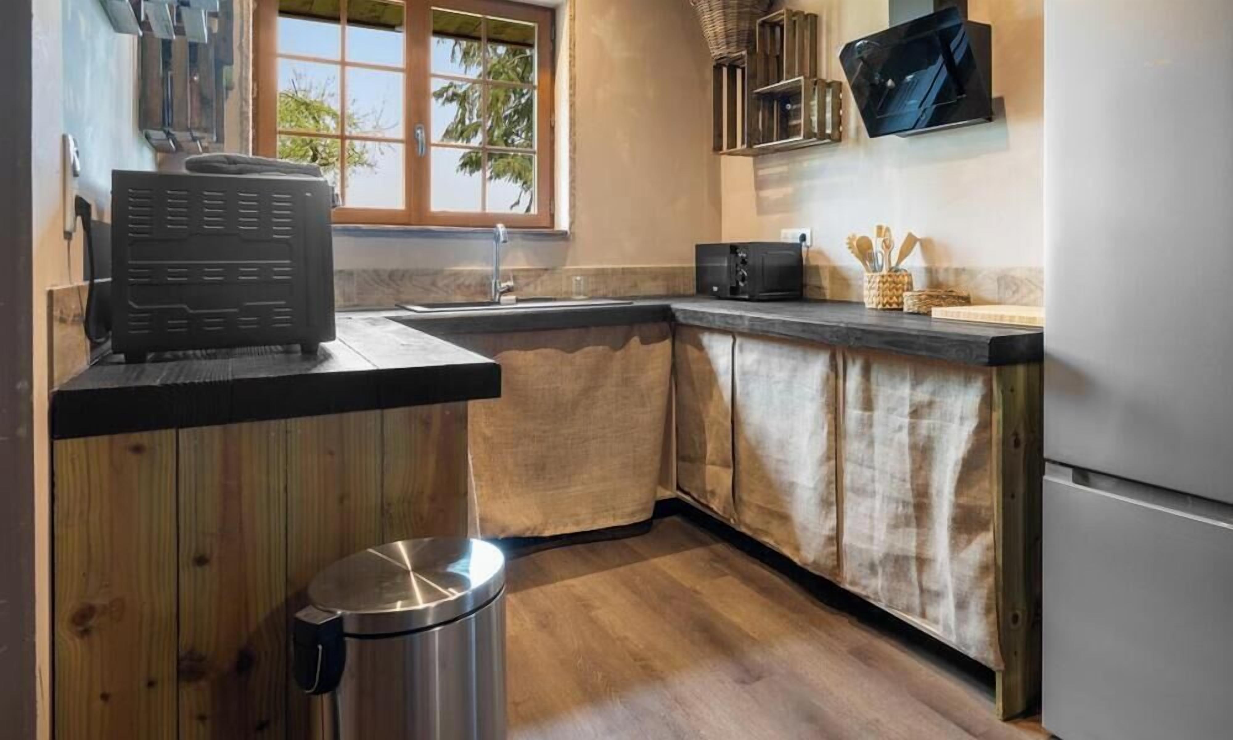 A compact kitchen with wooden cabinets, a stainless steel trash bin, a microwave, and a window with a view of greenery.