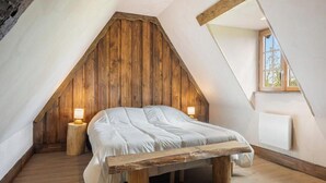 A loft bedroom with a sloped ceiling, wooden beams, a bed with white bedding, two bedside lamps, and a window with a view. - Gîtes de France® - Hygge (SAUMONT LA POTERIE)