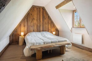 Un dormitorio tipo loft con techo inclinado, vigas de madera, una cama con ropa blanca, dos lámparas de noche y una ventana con vistas.