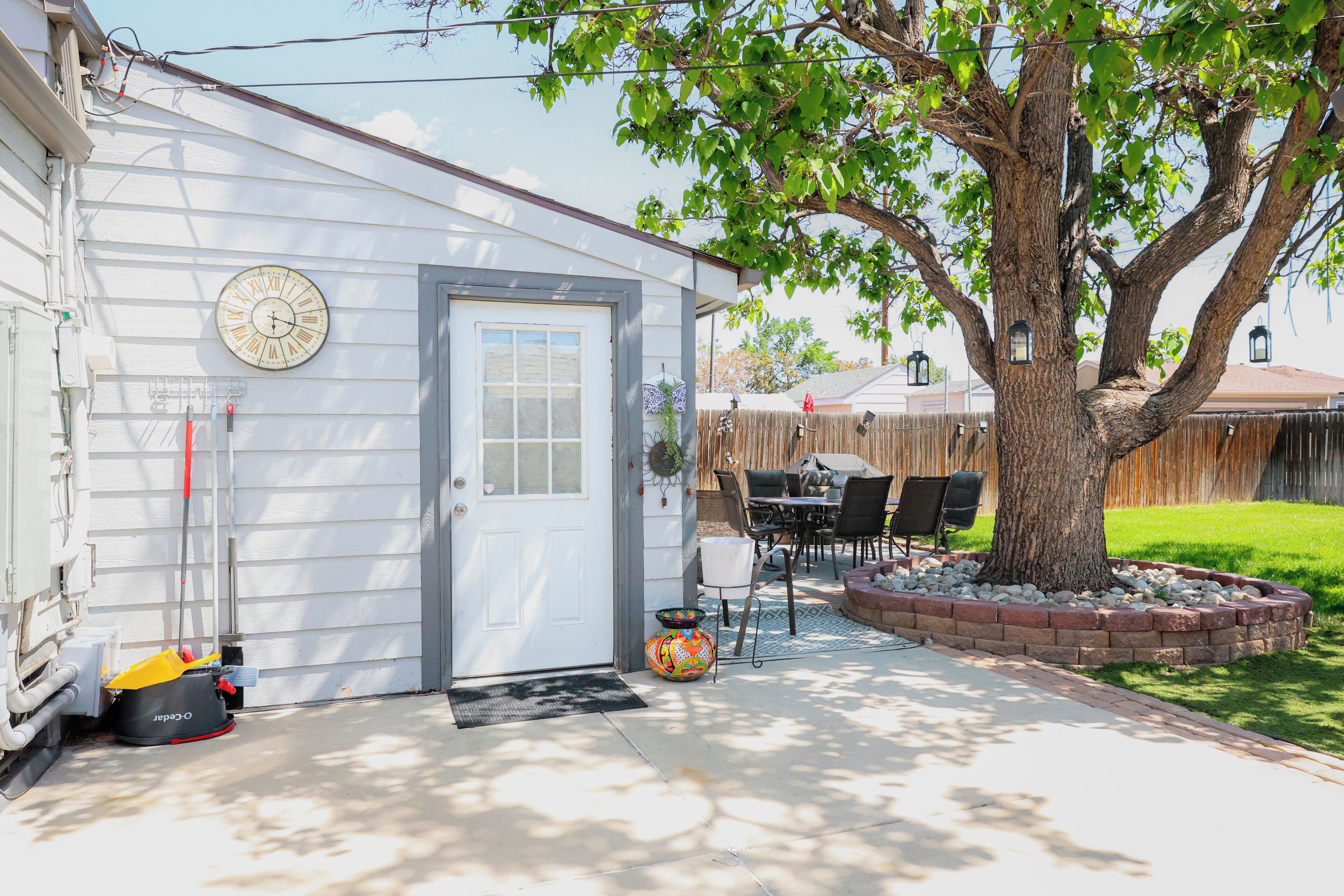 A house with a garage door, a tree, a patio with a table and chairs, and a clock on the wall.