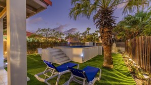Un giardino con piscina, due poltrone da relax, una palma e una recinzione in legno.