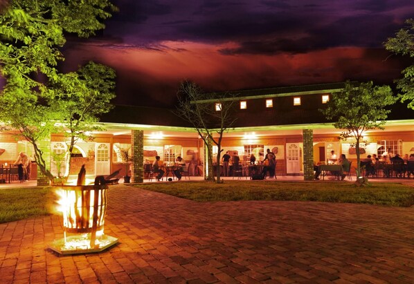 An outdoor dining area with brick flooring, a central fire feature, and a building with multiple windows and doors in the background. - Damara Mopane Lodge (Khorixas)
