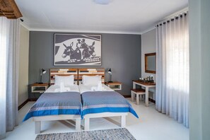 A double bed with a patterned bedspread, two bedside tables with lamps, a small desk with a mirror, and a framed artwork on the wall. - Damara Mopane Lodge (Khorixas)