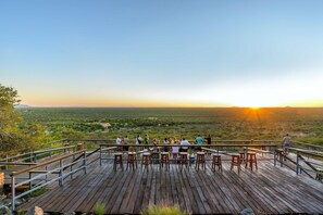 Una terraza de madera con mesas y sillas que da a un paisaje extenso al atardecer.
