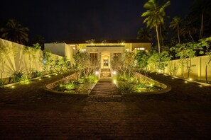 A nighttime view of a building with a central entrance, surrounded by a landscaped garden with pathways and palm trees. - Kayaam House (Rekawa)