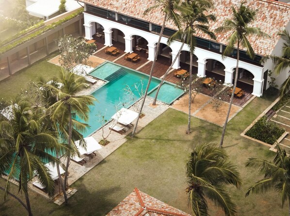 An aerial view of a resort with a swimming pool, palm trees, and a covered patio area.