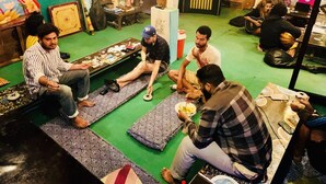 Um grupo de pessoas sentadas em esteiras estampadas em um quarto com paredes verdes, uma televisão e diversos objetos nas prateleiras.