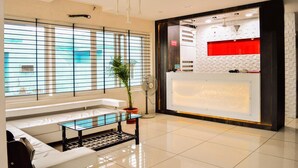 Um saguão de hotel com balcão de recepção, área de estar com sofá e mesa de centro, e grandes janelas com persianas.