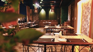 A restaurant with multiple tables and chairs, dim lighting, and hanging plants. - Hotel Rion (Udaipur)