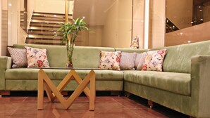 A green sectional sofa with floral cushions, a glass vase with green plants, and a geometric coffee table. - Hotel Rion (Udaipur)