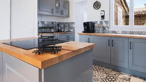 A modern kitchen with grey cabinets, a wooden countertop, a coffee machine, and a window with a view of a stone building. - Gîtes de France® - L'Orée D'Etretat (LA POTERIE CAP D'ANTIFER)