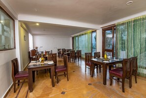 A spacious dining area with multiple wooden tables and chairs, large windows with green curtains, and a tiled floor.