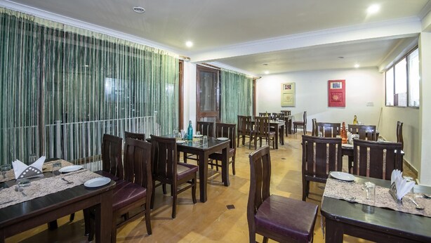 A dining area with wooden tables and chairs, a patterned floor, and a view through a window with curtains. - Xomotel Bungalows Krishanya (Nainital)