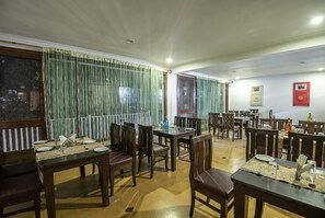 A dining area with multiple tables and chairs, a window with curtains, and a wall with framed pictures. - Xomotel Bungalows Krishanya (Nainital)