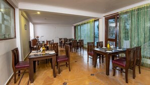 A spacious dining area with multiple wooden tables and chairs, large windows with green curtains, and a tiled floor. - Xomotel Bungalows Krishanya (Nainital)