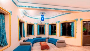 A room with a curved blue sofa, a small table, and large windows with curtains. - The Udai Bagh (Māvli)