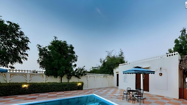 A swimming pool with a tiled deck, a covered seating area, and a white wall with decorative elements. - The Udai Bagh (Māvli)