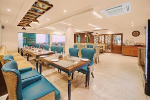 A dining area with multiple tables and chairs, a kitchen area, and a large window with a view. - Hotel Green One Earth (Yamkeshwar)