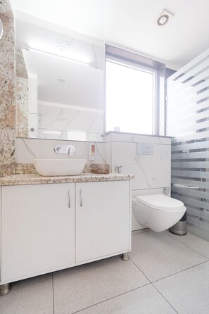 A modern bathroom with a white vanity, a large mirror, a toilet, and a window with blinds. - Hotel Green One Earth (Yamkeshwar)