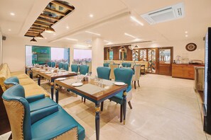 A dining area with multiple tables and chairs, a kitchen area with a refrigerator, and a view through the window. - Hotel Green One Earth (Yamkeshwar)